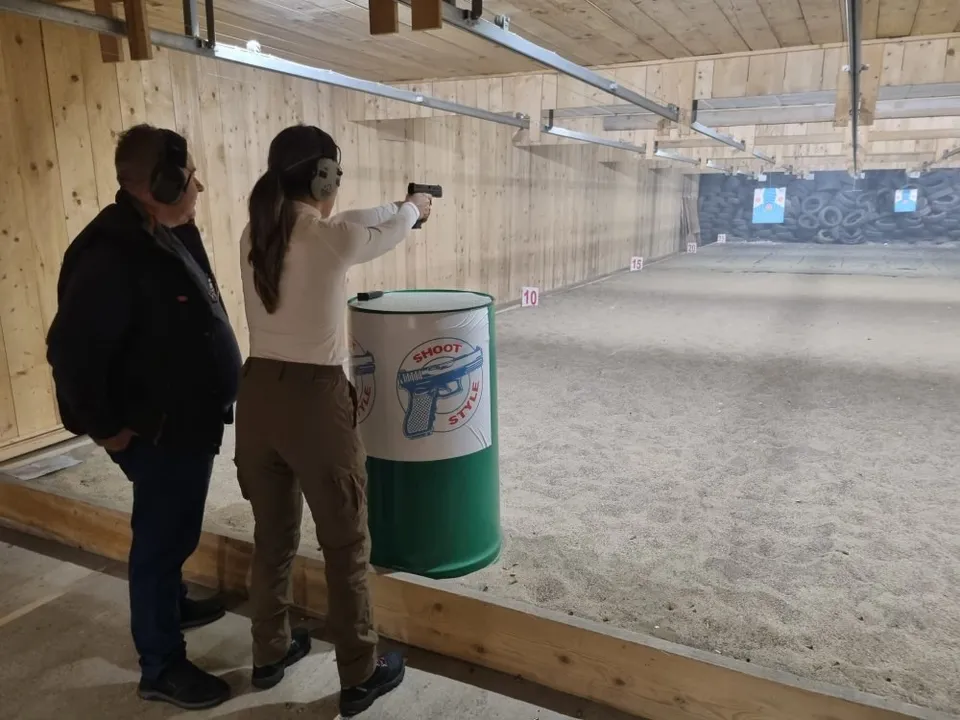 O femeie cu părul lung și o cămașă albă țintește un pistol într-un poligon de tragere, în timp ce un bărbat o privește de lângă ea.