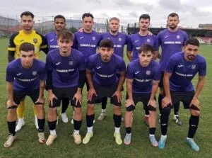 O echipă de fotbaliști îmbrăcați în echipamente albastre și galben posedă pentru o fotografie de grup pe un teren de fotbal sub un cer înnorat.