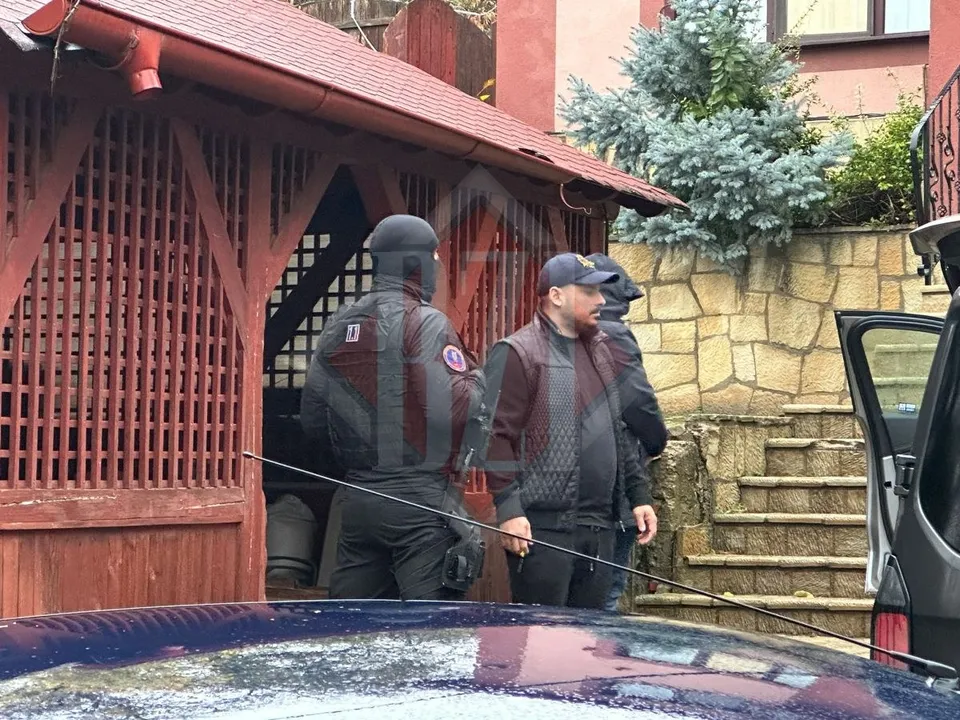 Oameni în echipamente tactice și civili stau în fața unei structuri de lemn cu grilaje, lângă o mașină și scări de piatră.