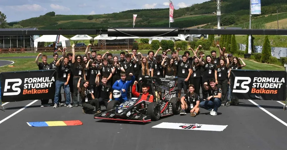 Un grup de studenți, majoritatea în tricouri negre, pozează în jurul unui monopost de curse negru, pe o pistă de asfalt, cu bannere "Formula Student Balkans" în fundal.