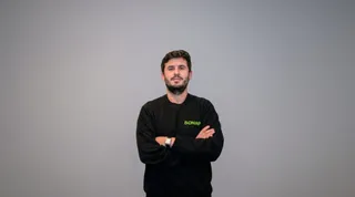 Un bărbat cu barbă și păr scurt, purtând un hanorac negru cu un logo verde, stă cu brațele încrucișate în fața unui fundal gri neutru.