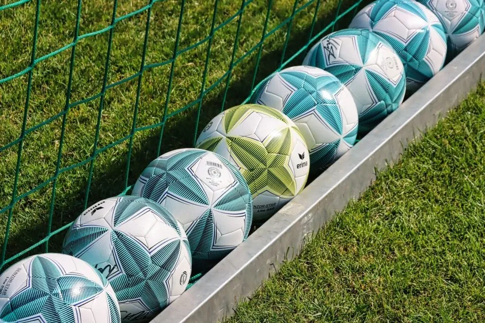 Mai multe mingi de fotbal colorate sunt așezate într-un suport metalic pe un teren de iarbă lângă o plasă verde.