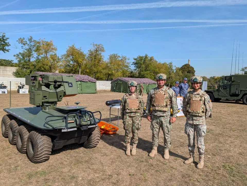 Trei persoane în uniforme militare de camuflaj stau lângă un vehicul militar mic cu roți și o platformă de echipament pe un câmp deschis.