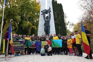 Un grup de oameni stă în fața unui monument înalt, ținând diverse steaguri și un banner colorat pe un teren pavat.