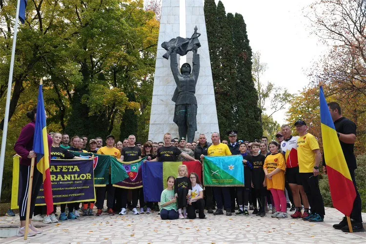 Un grup de oameni stă în fața unui monument înalt, ținând diverse steaguri și un banner colorat pe un teren pavat.