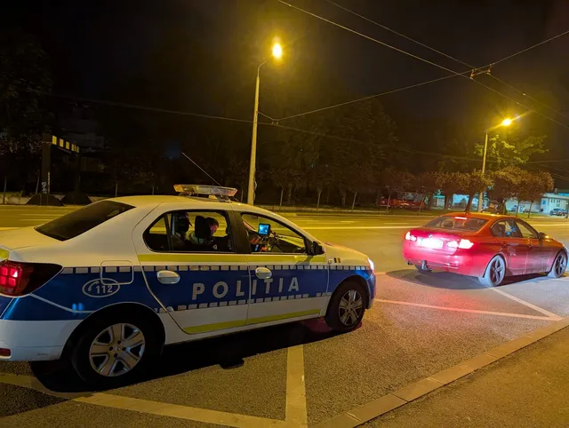 O mașină de poliție albă și albastră stă pe un drum asfaltat lângă o mașină roșie sub lumini de stradă noaptea.