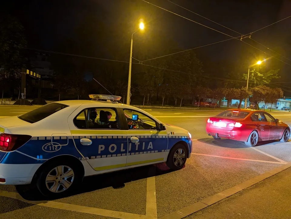 O mașină de poliție albă și albastră stă pe un drum asfaltat lângă o mașină roșie sub lumini de stradă noaptea.