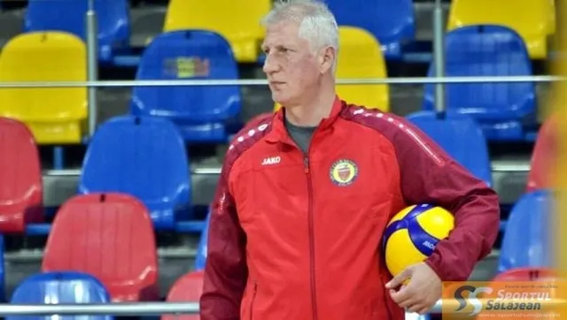 Un bărbat cu părul scurt și cămășă sportivă roșie ține o minge de volei galbenă și albastră în fața unor scaune colorate.