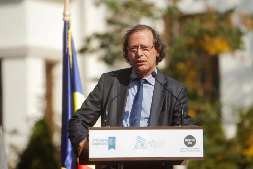 Un bărbat cu ochelari și sacou întunecat stă în spatele unui podium alb cu logouri, în fața unui steag albastru și galben.