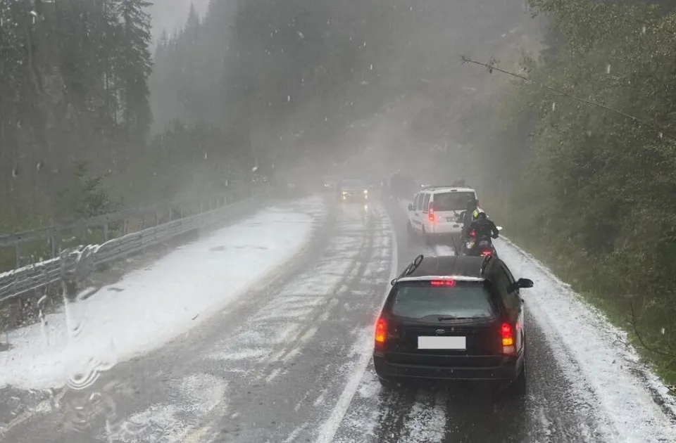 Mașini circulă pe un drum montan asfaltat, acoperit parțial de zăpadă, în condiții de ceață și precipitații.
