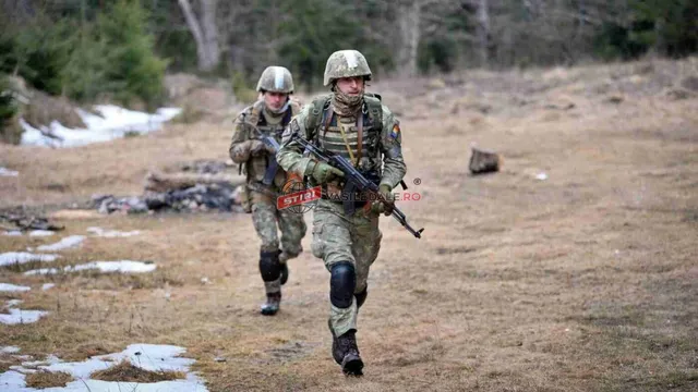 Doi soldați în uniforme de camuflaj și căști de protecție aleargă pe un teren împădurit cu zone cu resturi de zăpadă.