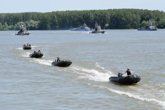 Mai multe bărci cu motor mici și nave militare navighează pe un fluviu larg, lăsând urme de spumă albă pe apă.