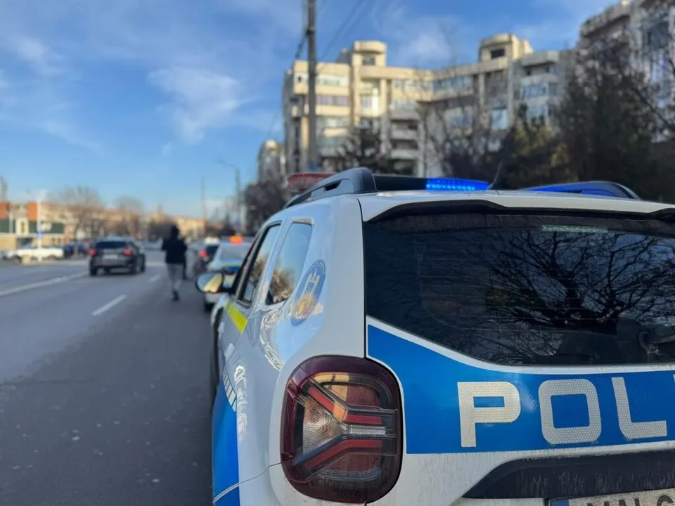 Partea din spate a unui vehicul alb cu marcaje albastre și textul POL, parcat pe marginea unui drum asfaltat cu clădiri și mașini în fundal.