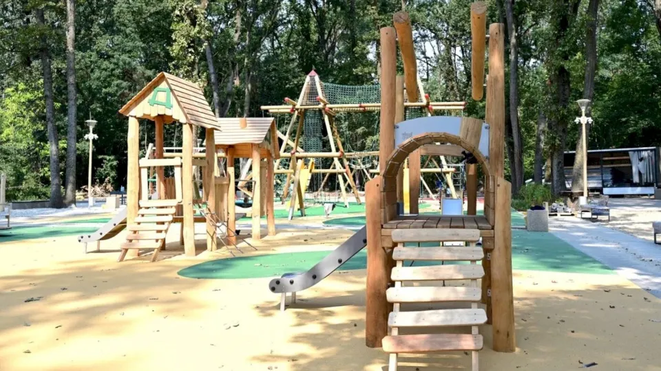 Echipamente de joacă din lemn cu tobogane și scări sunt instalate pe un teren cu suprafețe de protecție colorate într-o zonă împădurită.
