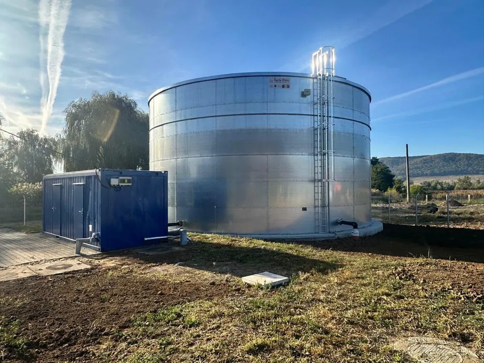 Un rezervor cilindric metalic de culoare argintie este situat lângă un container albastru pe un teren cu iarbă și pământ sub un cer senin.