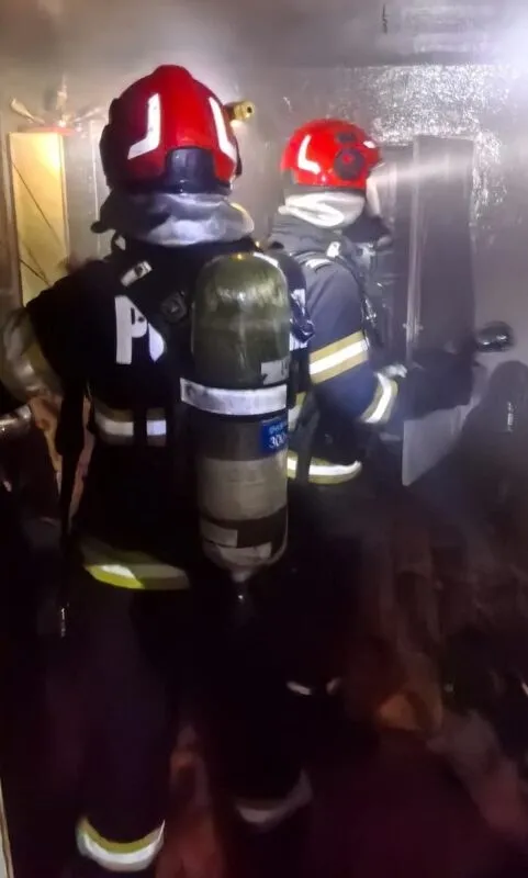 Doi pompieri cu echipament de protecție, casci roșii și butelii de oxigen, sunt poziționați într-un spațiu întunecat.