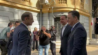 Patru bărbați în costume purtând ochelari stau în picioare pe o stradă lângă un grup de oameni care fac fotografii.