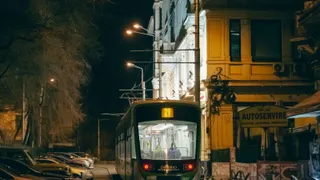 Un tramvai circulă pe un drum urban noaptea, lângă clădiri iluminat de felinare și de luminile vehiculului.