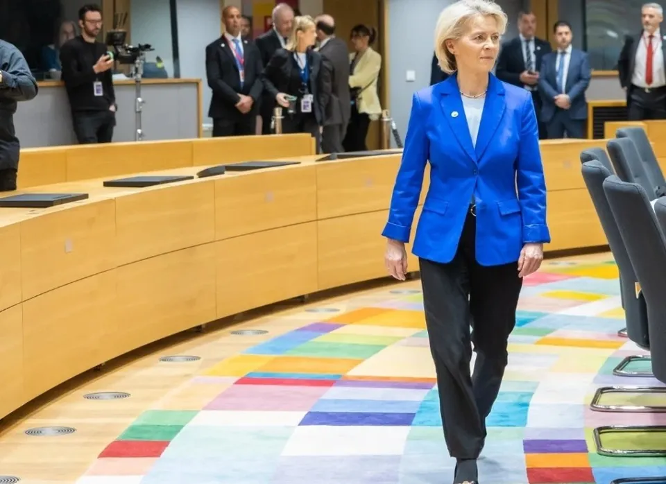 O femeie cu părul scurt și bluză albă poartă un sacou albastru și pantaloni negri, mergând pe un covor colorat într-o sală de conferințe.