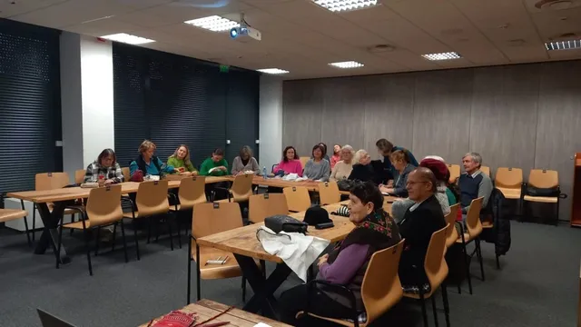 Un grup de persoane stă așezat la mese în interiorul unei săli de conferințe cu pereți gri și scaune de culoare portocalie.