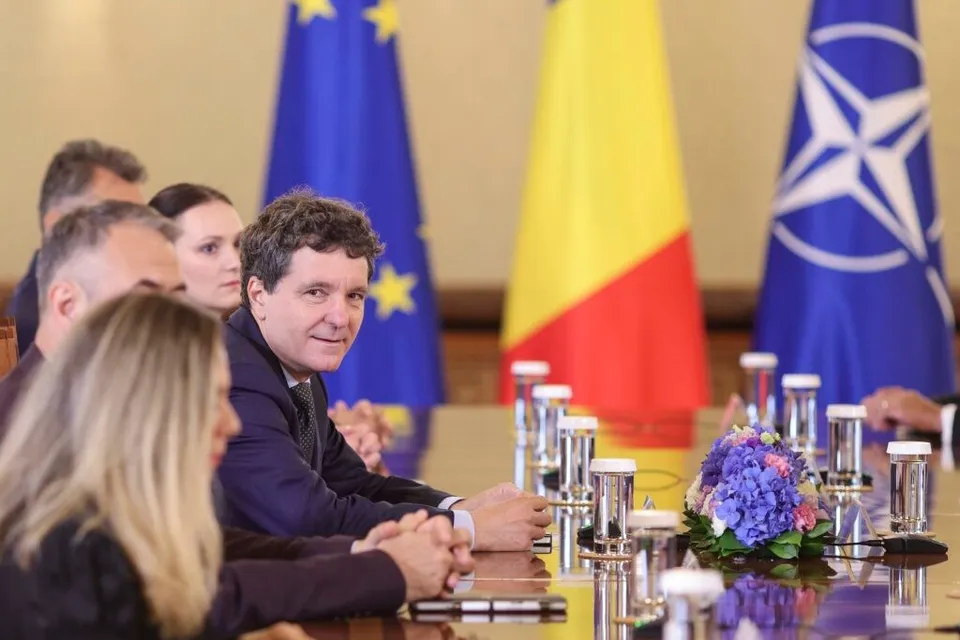 Un bărbat cu părul creț și sacou închis se întoarce spre cameră stând la o masă de conferință cu alte persoane și steaguri în fundal.