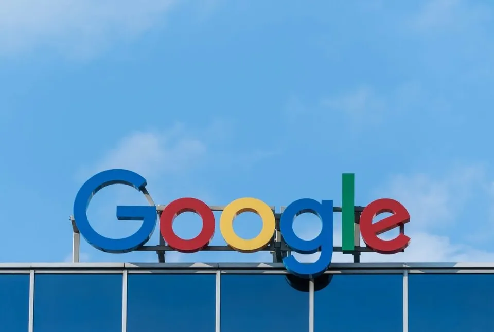 Un logo colorat Google este montat pe partea superioară a unei clădiri cu fațadă de sticlă albastră sub un cer senin.
