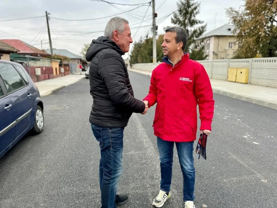 Un bărbat într-o jachetă neagră și pantaloni de jeanș strânge mâna unui bărbat într-o jachetă roșie și pantaloni de jeanș pe un drum asfaltat.