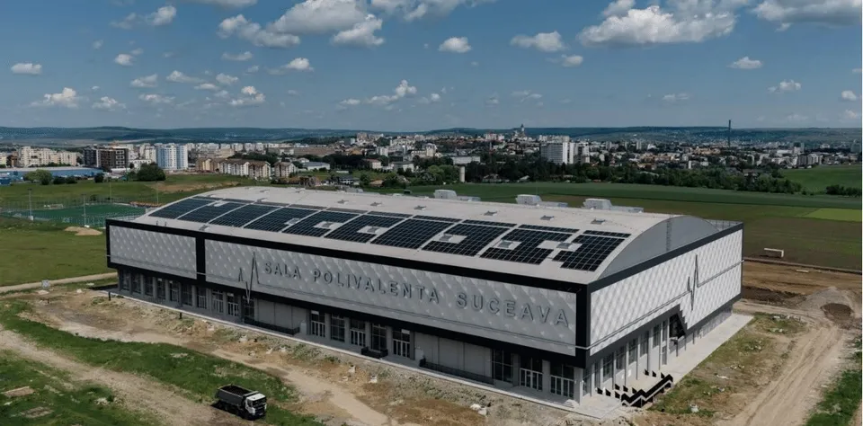 O clădire mare, rectangulară, cu panouri solare pe acoperiș, este situată pe un teren de culoare verde sub un cer cu nori.