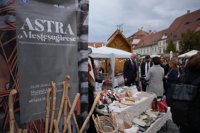 Un banner gri cu textul ASTRA Meșteșugăreșe stă în prim-plan lângă obiecte din lemn, în timp ce oameni vizitează tarabe cu diverse produse în fața unor clădiri.