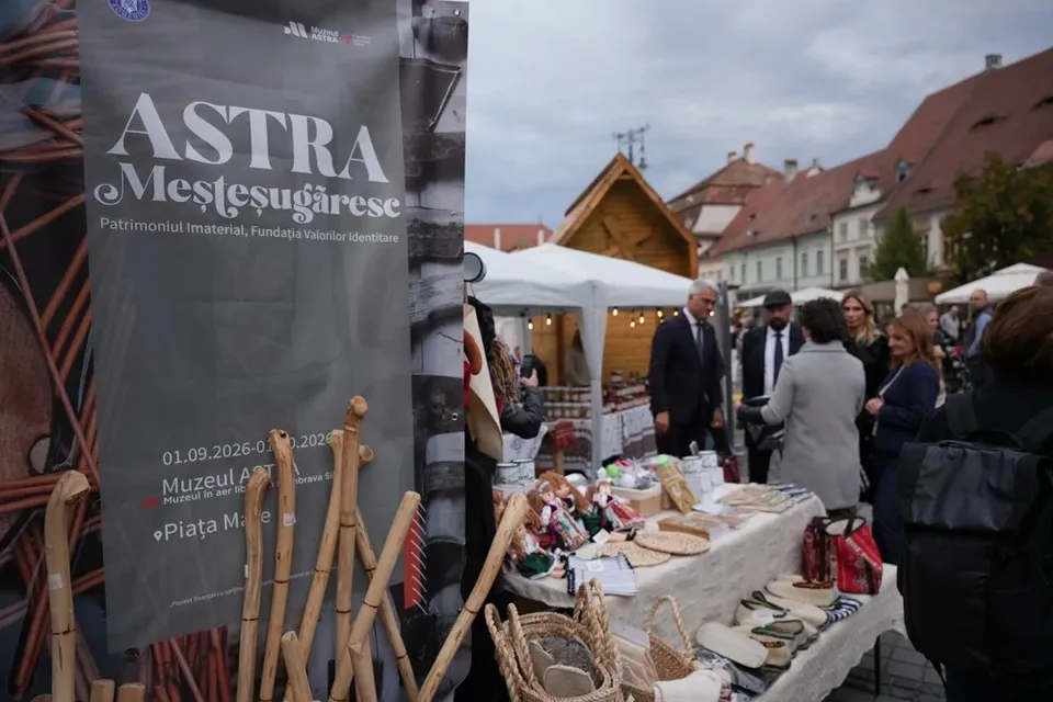 Un banner gri cu textul ASTRA Meșteșugăreșe stă în prim-plan lângă obiecte din lemn, în timp ce oameni vizitează tarabe cu diverse produse în fața unor clădiri.