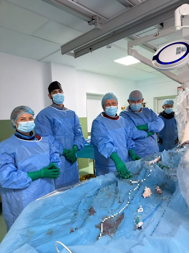 Cinci persoane îmbrăcate în halate medicale albastre și mănuși verzi stau în jurul unui pat de operație acoperit cu un drapel steril albastru într-o sală de spital.