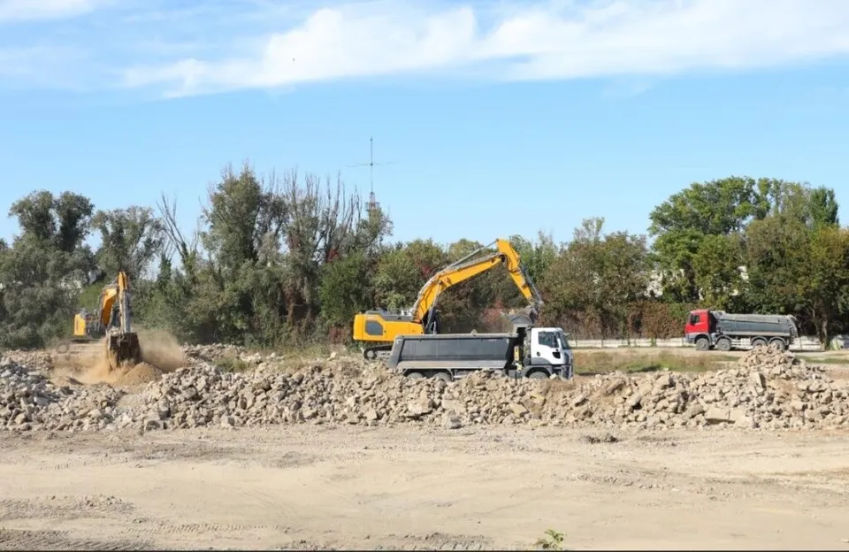 Două excavatoare galbene și un camion alb lucrează pe un teren de construcție cu grămezi de pământ și copaci în fundal sub un cer senin.