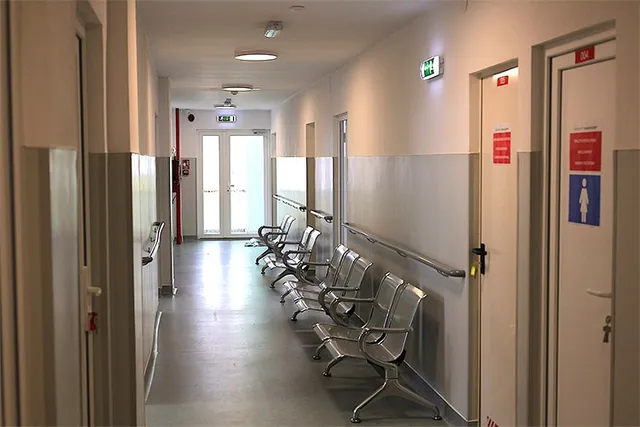 Un hol de spital cu un rând de scaune metalice de așteptare dispuse de-a lungul unui perete alb.