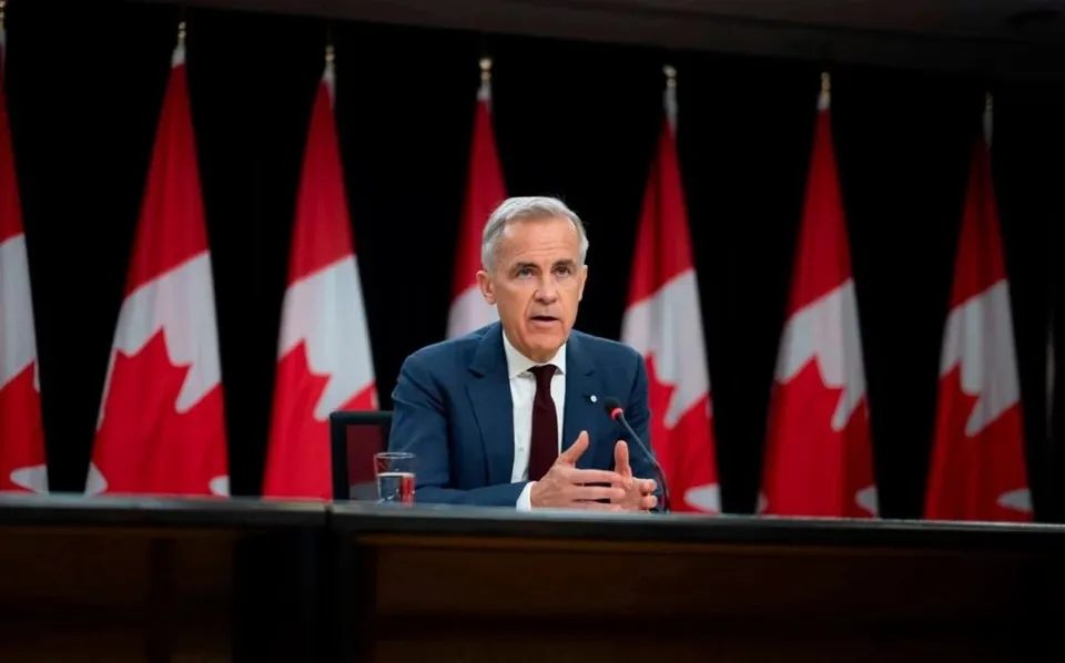 Un bărbat cu părul cărunt, purtând costum albastru și cravată închisă, stă la o masă în fața mai multor steaguri canadiene.