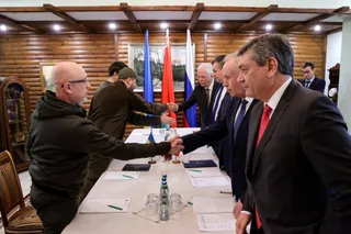 Mai mulți bărbați stau în jurul unei mese albe și dau mâna în timp ce steaguri sunt așezate în fundal.