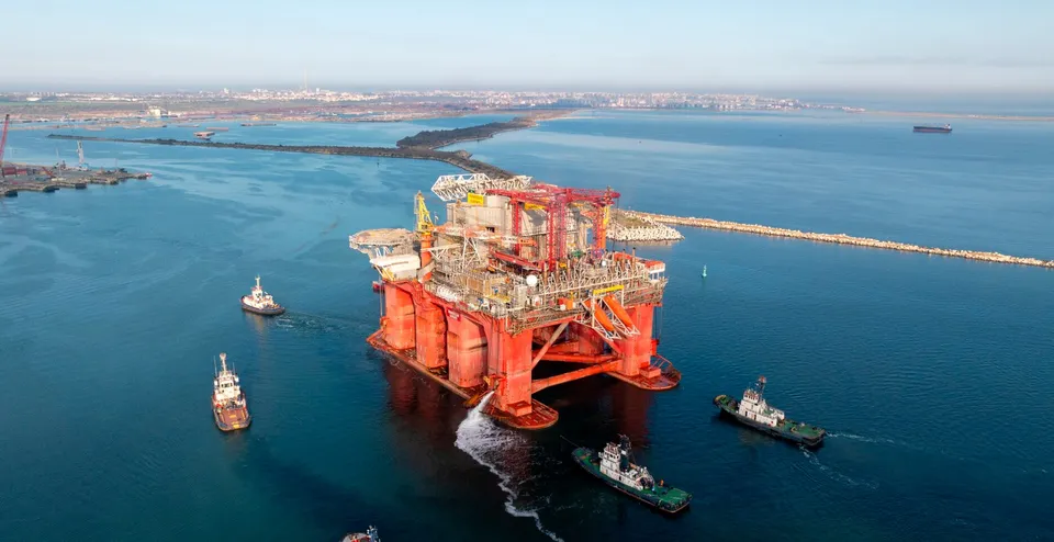 O platformă petrolieră de culoare portocalie este înconjurată de patru nave mici pe un lac sau o mare cu apă albastră.