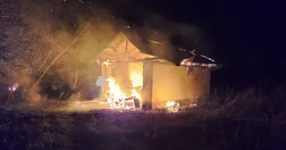 O clădire mică cu acoperiș în pante arde intens noaptea, emanând flăcări și fum în apropierea unor vegetații întunecate.