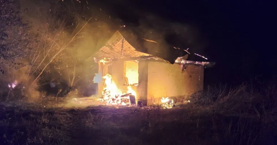 O clădire mică cu acoperiș în pante arde intens noaptea, emanând flăcări și fum în apropierea unor vegetații întunecate.