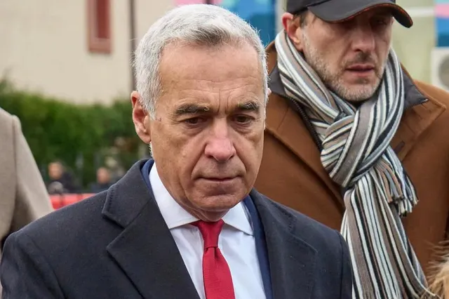 Un bărbat cu părul cărunt, purtând sacou închis la culoare, cămașă albă și cravată roșie, privește în jos lângă un alt bărbat cu șapcă și eșarfă în dungi.