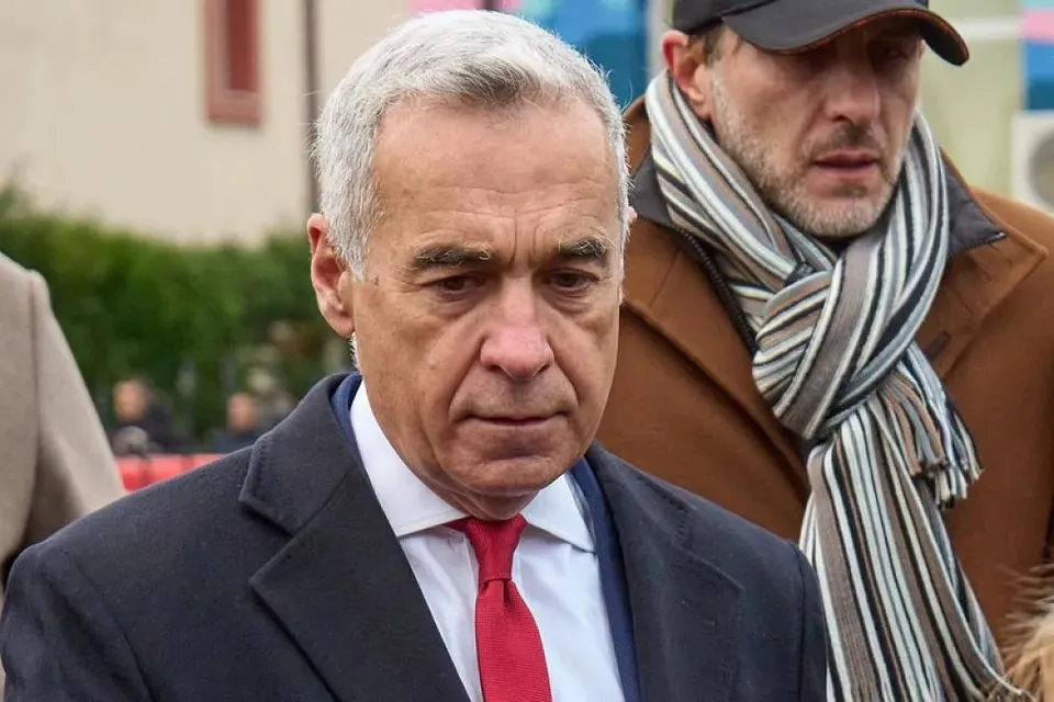 Un bărbat cu părul cărunt, purtând sacou închis la culoare, cămașă albă și cravată roșie, privește în jos lângă un alt bărbat cu șapcă și eșarfă în dungi.