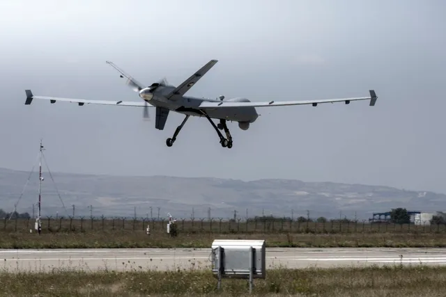 Un avion fără pilot de dimensiuni mari zboară deasupra unei piste de aterizare lângă un teren cu iarbă și vegetație scundă sub un cer înnorat.