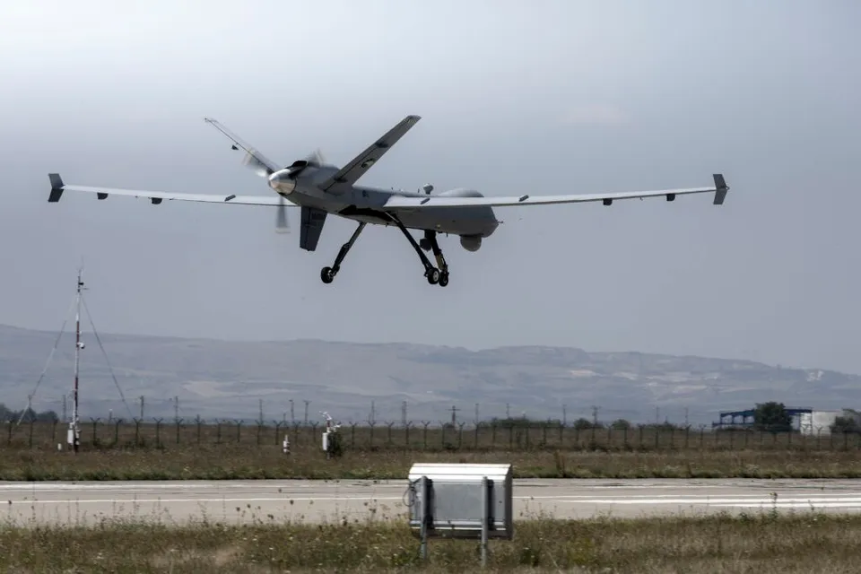 Un avion fără pilot de dimensiuni mari zboară deasupra unei piste de aterizare lângă un teren cu iarbă și vegetație scundă sub un cer înnorat.