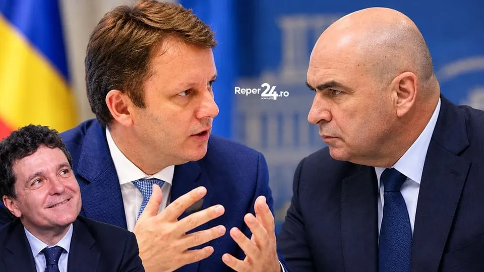 Un bărbat cu părul castaniu într-un costum albastru vorbește gesticulând către un bărbat chel cu costum închis și cravată albastră.