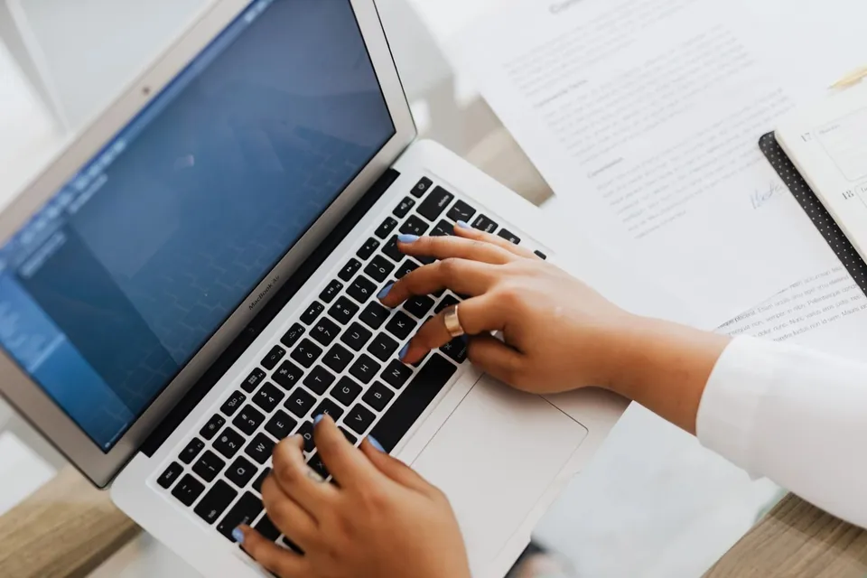 O persoană cu unghiile vopsite în albastru tastează pe un laptop alb aflat lângă documente pe o masă de lucru.