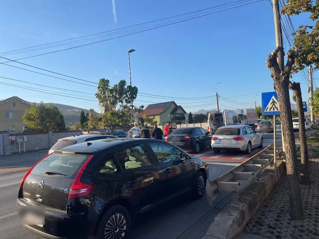 Două autoturisme negre și un sedan alb sunt parcate pe marginea unui drum asfaltat sub un cer senin.