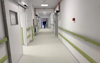 Un hol de spital cu pereți albi, coridoare lungi, balustrade verzi și două persoane mergând în depărtare.