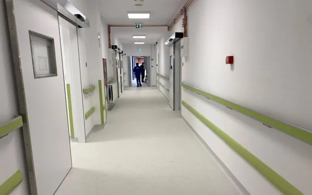 Un hol de spital cu pereți albi, coridoare lungi, balustrade verzi și două persoane mergând în depărtare.