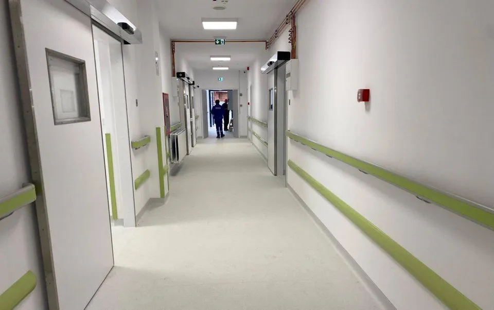Un hol de spital cu pereți albi, coridoare lungi, balustrade verzi și două persoane mergând în depărtare.