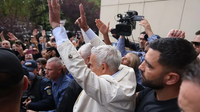 Un grup de persoane stă într-o mulțime, în timp ce un bărbat cu părul alb ridică brațul drept în sus.