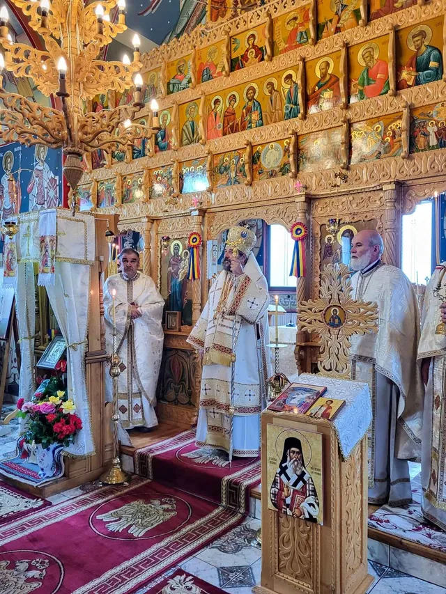 Trei bărbați în haine liturgice albe stau în interiorul unei biserici lângă un iconostas decorat cu picturi religioase și un candelabru de aur.